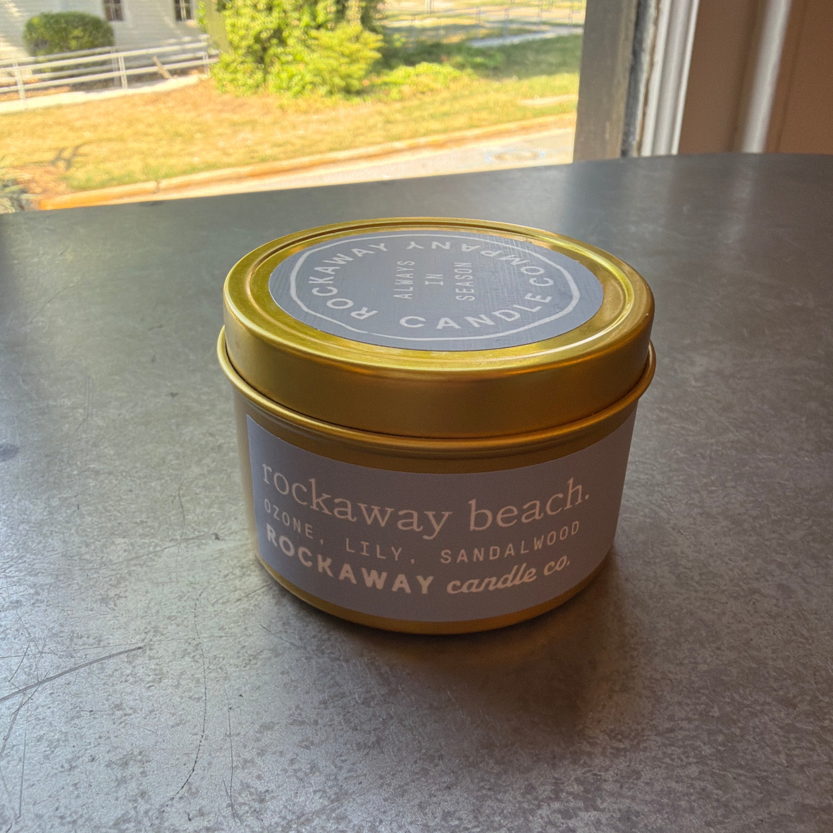 Rockaway Travel Candle, 4 oz., Beach in a gold tin with a lavender label sitting on a gray surface by a window with outdoor greenery in the background, perfect for cozy decor or gifts from Knotty and Board.