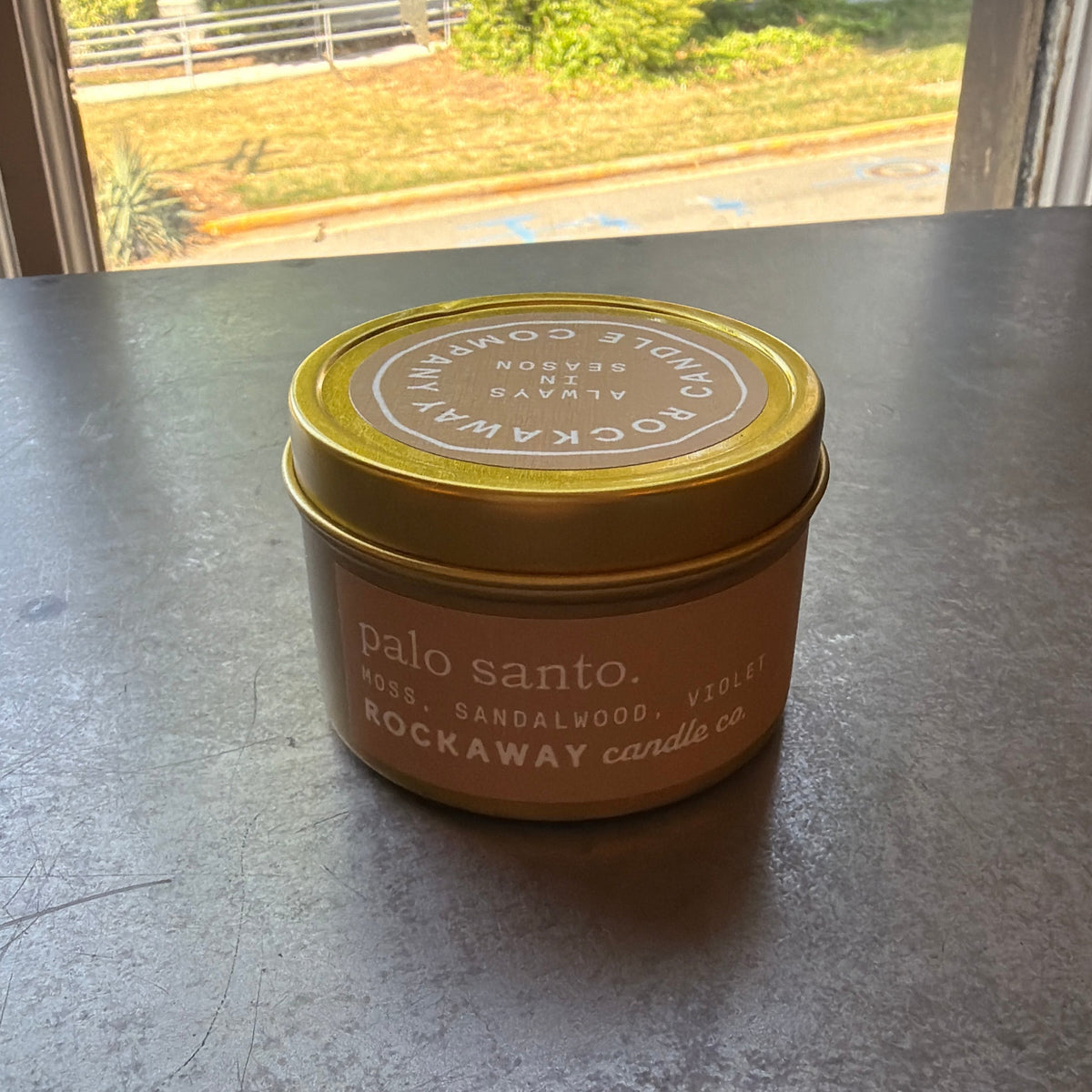 Rockaway Travel Candle 4oz Palo Santo in a small brown tin with a gold lid, placed on a dark surface with natural light from a window nearby, perfect for home decor and gifts.