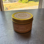 Rockaway Travel Candle 4oz Palo Santo in a small brown tin with a gold lid, placed on a dark surface with natural light from a window nearby, perfect for home decor and gifts. 1