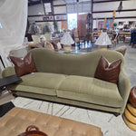 An olive green velvet loveseat sofa with two brown leather accent pillows, positioned in a furniture showroom with a tufted coffee table and wooden stools in the foreground. 2