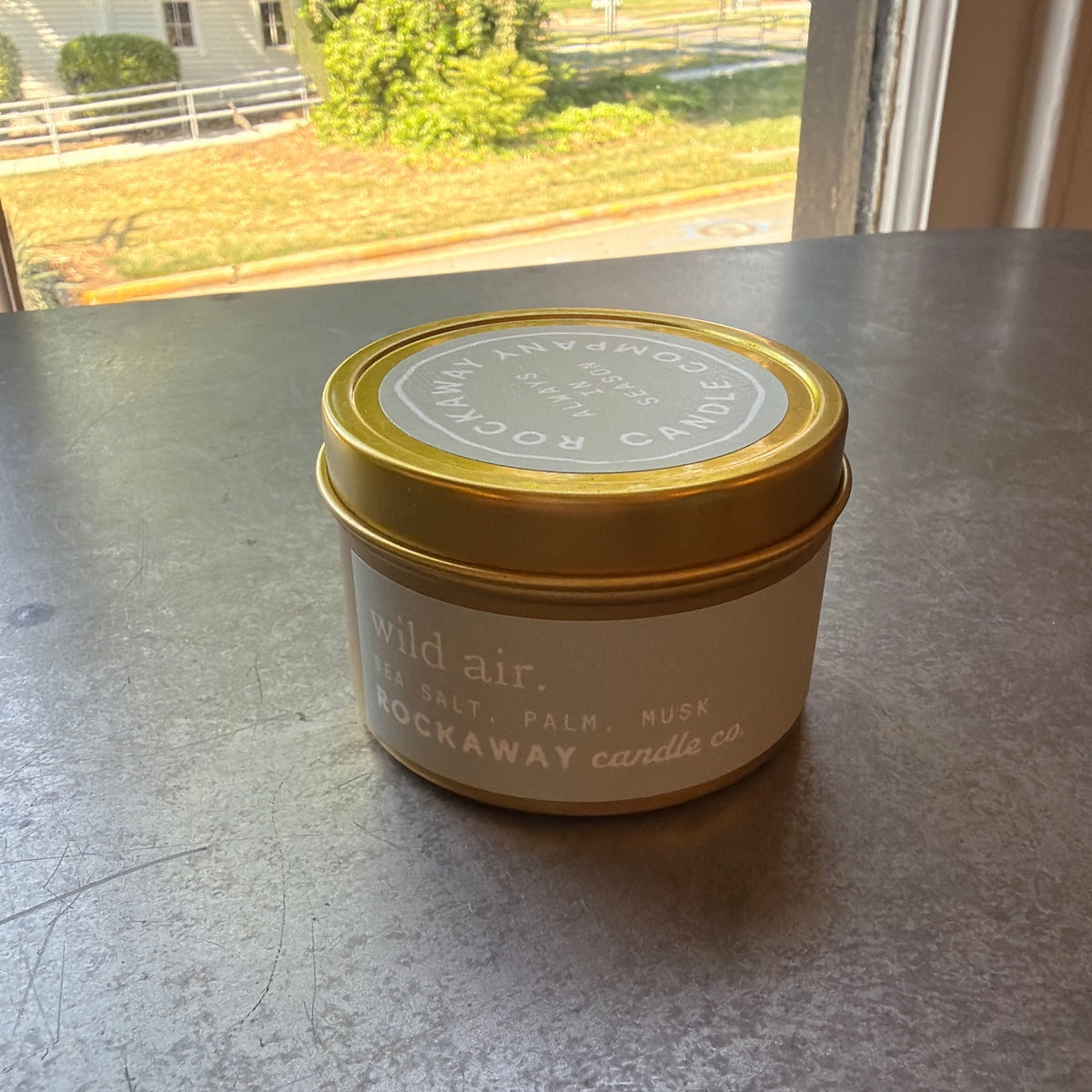 Rockaway Travel Candle 4oz Wild Air with gold metal lid, white label featuring natural scents of salt, palm, and musk, displayed on a kitchen countertop by a sunlit window, decor and gift idea from Knotty and Board, Every Room Needs A Little Knotty and Board.