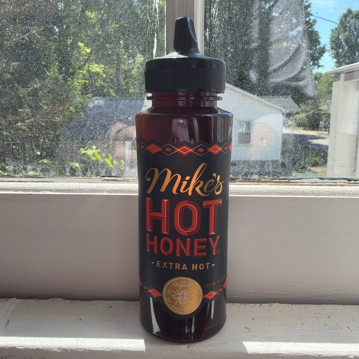 Mike's Hot Honey Squeeze Bottle, Extra Hot, 12 oz. standing on a sunny windowsill with outdoor greenery backdrop, adding a spicy touch to kitchen and dining decor.