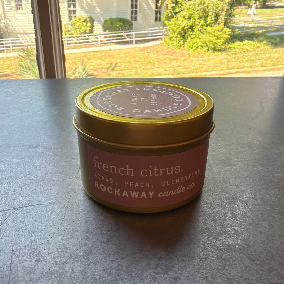 Rockaway Travel Candle 4oz French Citrus in a gold tin with pink label sitting on a windowsill with sunlight and outdoor greenery in the background, curated decor and gifts from Knotty and Board.