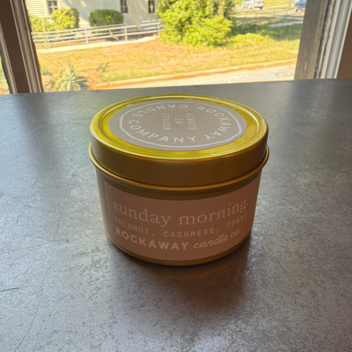 Rockaway Travel Candle 4oz Sunday Morning in a small tin with yellow lid, placed on a table near a sunlit window, perfect for home decor or gifts from Knotty and Board.