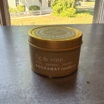 Rockaway Travel Candle 4oz Fig & Vine in a small gold tin with a lid, sitting on a dark surface near a sunny window with trees and a white fence visible outside. 1