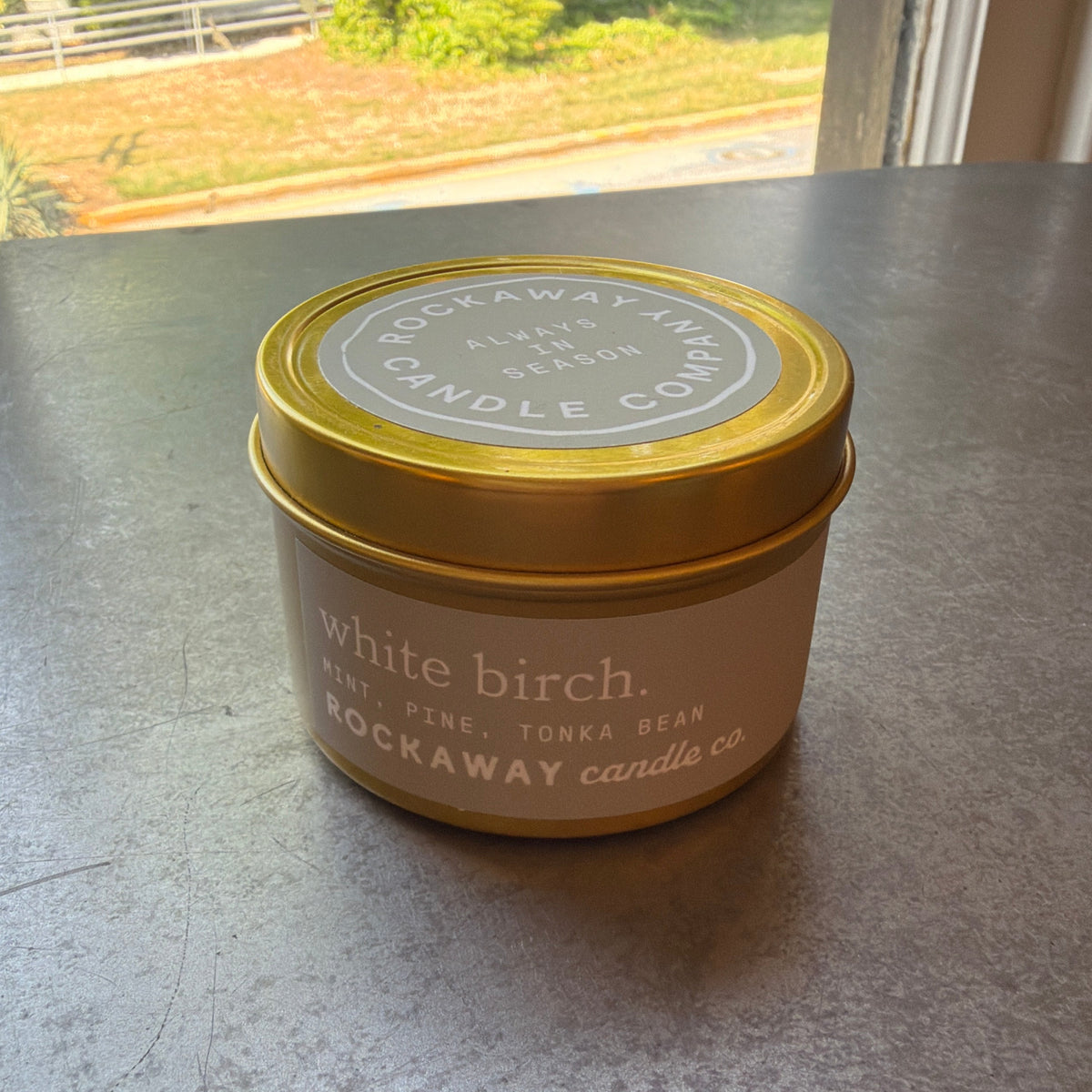 Rockaway Travel Candle 4oz White Birch with gold lid sitting on a dark surface near a window, showcasing elegant home decor and cozy ambiance from Knotty and Board for gifts and entertaining.
