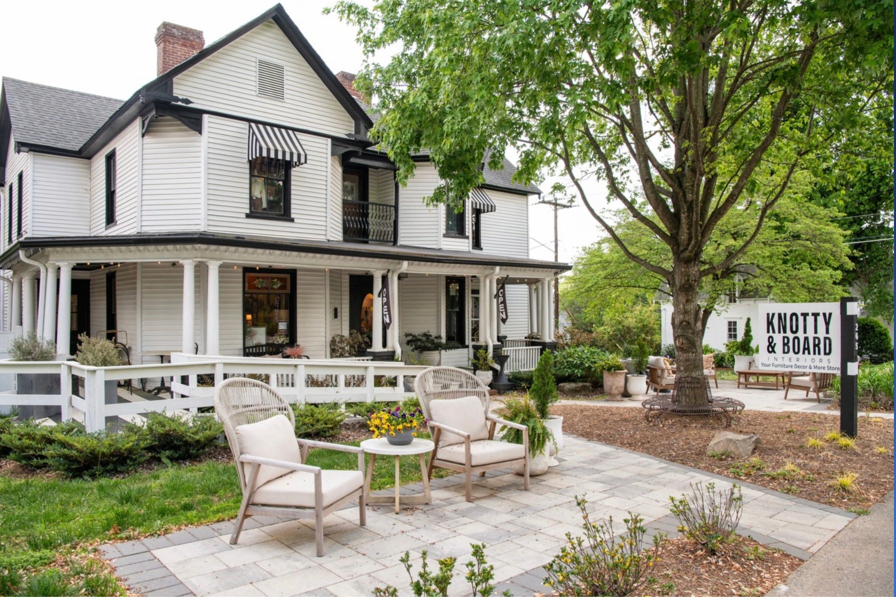 A charming white two-story house with a porch and black-striped awnings, featuring outdoor chairs and a table on a paved patio, next to a large tree and a sign for Knotty & Board decor and gifts.