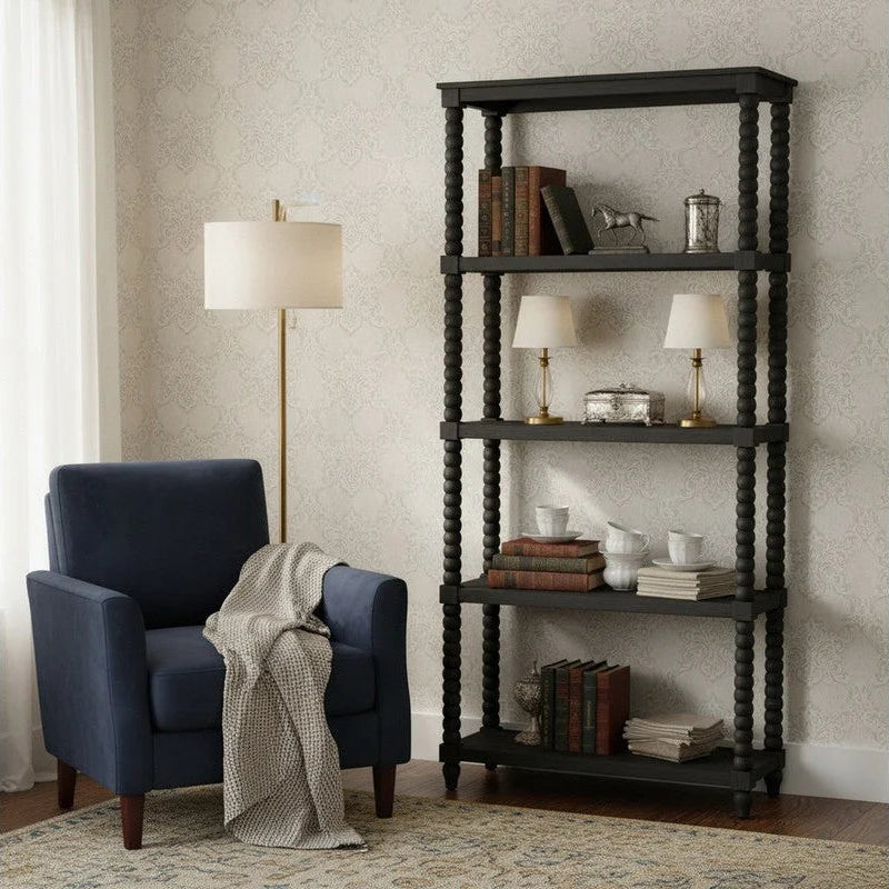 A tall black open bookcase with four shelves stands beside a blue armchair in a styled room. The shelves are decorated with books, small lamps, and decorative objects.