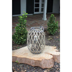 Grey Willow Lantern with Glass, Small, 16 in., displayed outdoors on a natural wood slab surrounded by garden plants, perfect for decorative candles and cozy outdoor entertaining. 1