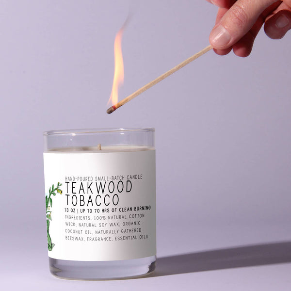 A hand holding a lit matchstick lighting a Just Bee Teakwood Tobacco - 7 oz candle in a clear glass container, showcased as a thoughtful gift and home decor item from Knotty and Board.