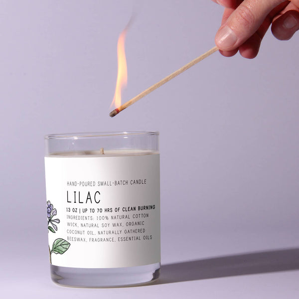 A hand holding a lit match above a Just Bee Lilac Candle, 7 oz, with a simple white label featuring lilac flower decor, perfect for home decor or gifts from Knotty and Board.