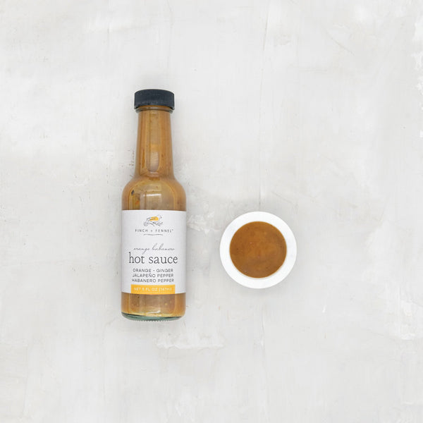 Bottle of Orange Habanero Hot Sauce, 5 oz next to a small bowl of the sauce, styled with a clean kitchen background for Knotty and Board kitchen and dining decor and gifts.