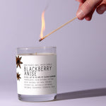 Hand lighting a Just Bee Blackberry Anise, 13 oz candle with a wooden wick in a clear glass jar, perfect for home decor, gifts, and cozy kitchen ambiance from Knotty and Board. 1