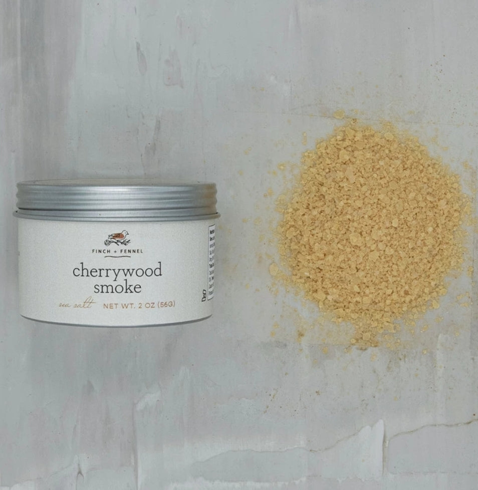 Finch & Fennel Cherrywood Smoked Sea Salt, 2 oz container next to a pile of light brown smoked sea salt on a kitchen surface, ideal for gifts and kitchen decor from Knotty and Board.