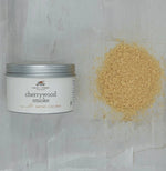 Finch & Fennel Cherrywood Smoked Sea Salt, 2 oz container next to a pile of light brown smoked sea salt on a kitchen surface, ideal for gifts and kitchen decor from Knotty and Board. 1