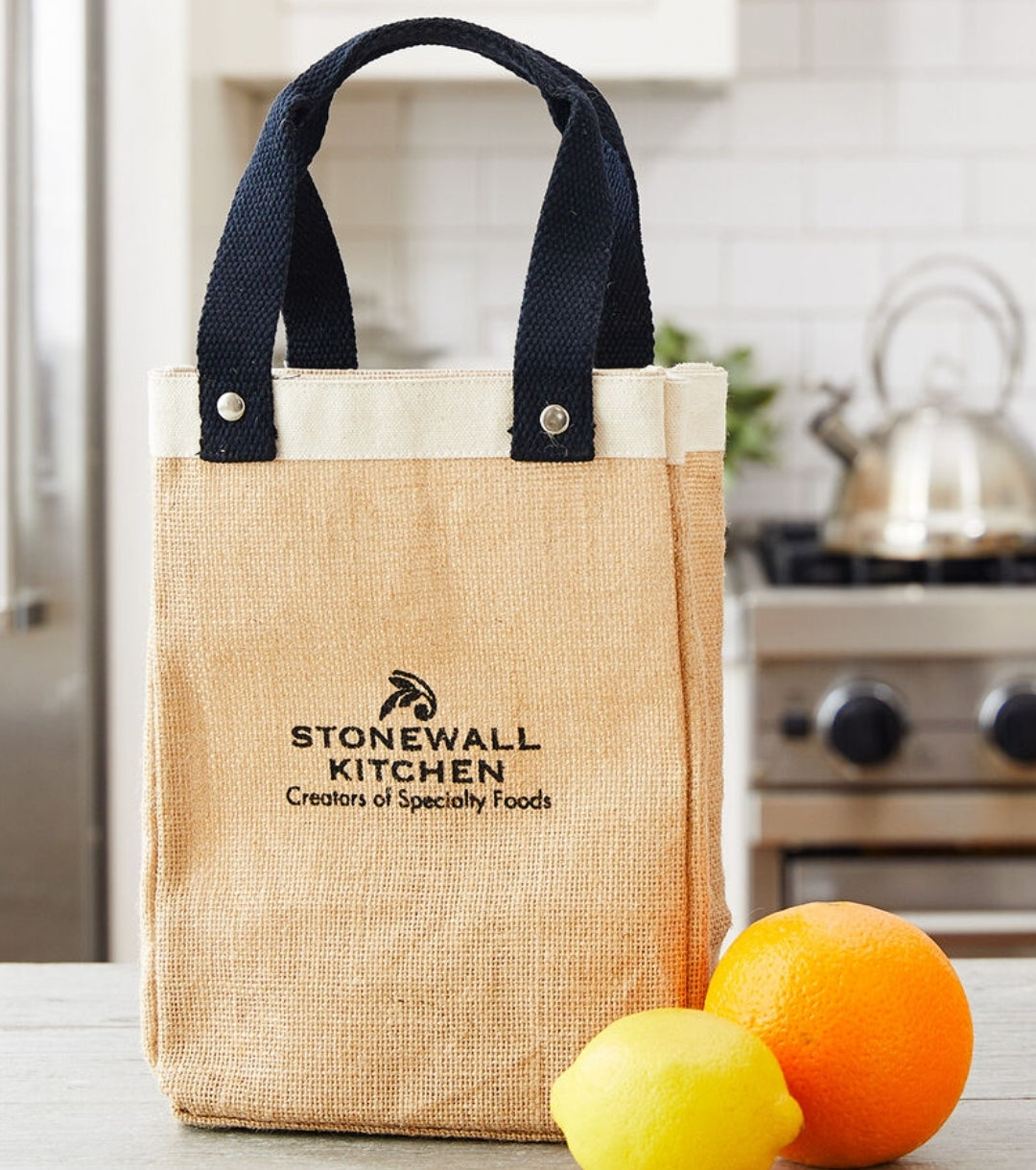 Stonewall Kitchen Farmers' Market Tote made of natural burlap with black sturdy handles, sitting on a kitchen counter near a lemon and orange, perfect for grocery or gift shopping.