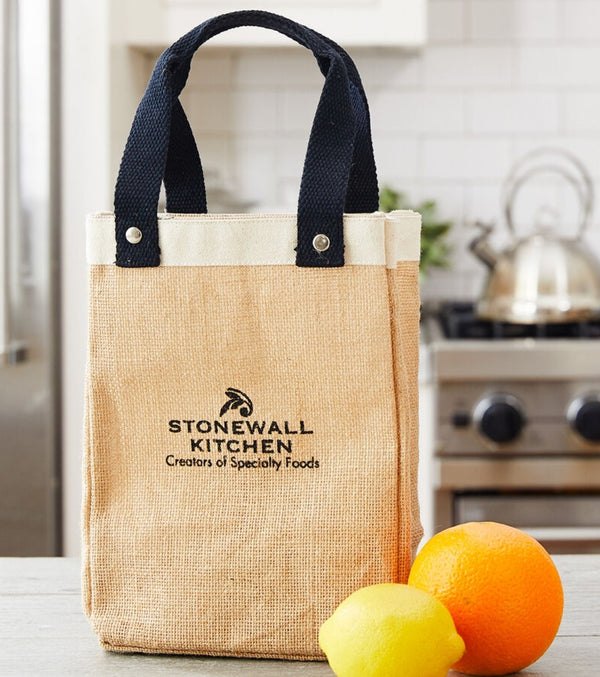 Stonewall Kitchen Farmers' Market Tote made of natural burlap with black sturdy handles, sitting on a kitchen counter near a lemon and orange, perfect for grocery or gift shopping.