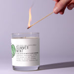 Hand lighting a Just Bee Summer Mint Candle - 7 oz with natural ingredients, placed on a light surface highlighting cozy decor and gift ambiance from Knotty and Board. 1