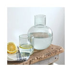Recycled Glass Bedside Carafe Set featuring a clear glass carafe and matching tumbler with water and lemon slice, styled on rustic wooden surface with a fresh lemon half nearby. 1