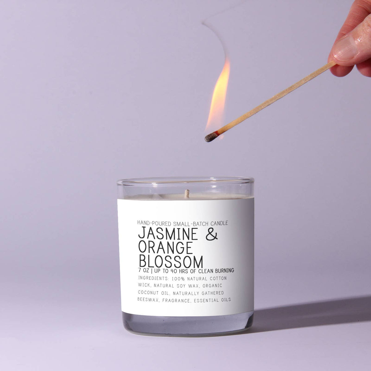 Hand lighting a Just Bee Jasmine and Orange Blossom Candle, 7 oz, in a clear glass jar with a white label against a pale background, perfect for home decor and gifts from Knotty and Board.