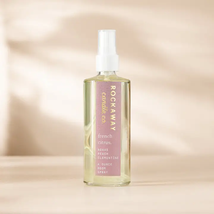 Rockaway French Citrus Room Spray in a clear spray bottle with a white nozzle, featuring a pink label, placed on a light wood surface with soft natural light and shadows in the background.