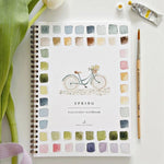 Watercolor Workbook, Spring featuring a spiral-bound cover with a painted bicycle and floral basket surrounded by colorful swatches, a paintbrush, and a tube of watercolor paint next to a tulip leaf on a white surface, offered by Knotty and Board with the tagline Every Room Needs A Little Knotty and Board. 1