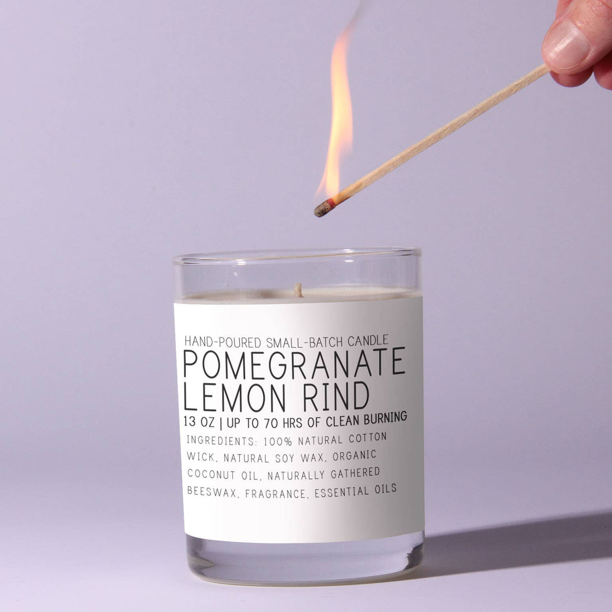 A hand-poured Just Bee Pomegranate & Lemon Rind Candle, 13 oz, with a natural cotton wick being lit by a match, ideal for decor and gifts from Knotty and Board.