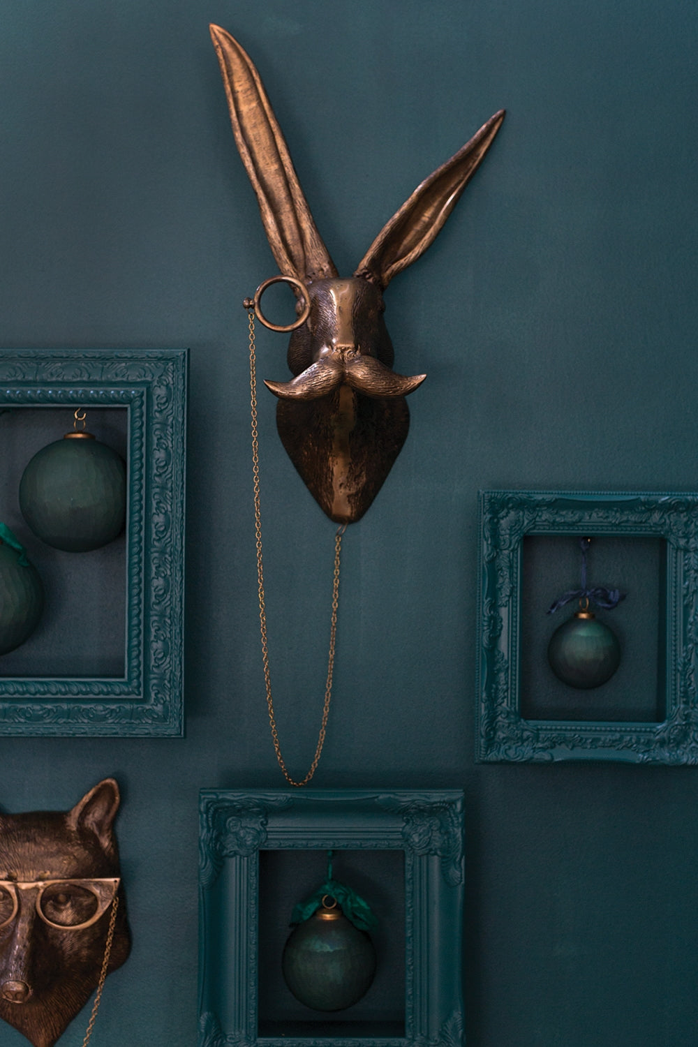 E&E Brass Wall Mount, Eric, features a whimsical brass hare head with a monocle and mustache, mounted on a deep teal wall accented with dark teal frames and ornaments, perfect for unique home decor from Knotty and Board. Every Room Needs A Little Knotty and Board.