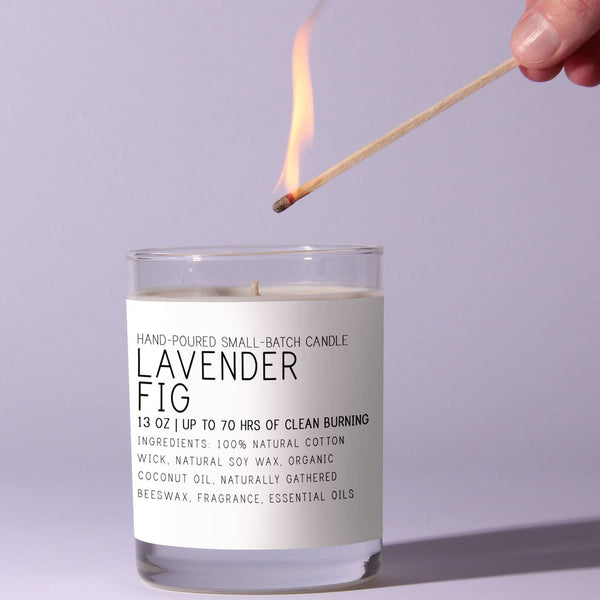 Person lighting a Just Bee Lavender Fig Candle - 7 oz with a wooden matchstick against a soft lavender background, showcasing a clean, minimalistic design perfect for home decor and gifts.