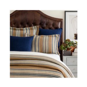 A cozy bedroom scene featuring the Heron Blanket, Full/Queen, Blue Bedding layered on a bed with striped and navy pillows, complemented by a leather headboard and bedside decor from Knotty and Board.