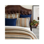 A cozy bedroom scene featuring the Heron Blanket, Full/Queen, Blue Bedding layered on a bed with striped and navy pillows, complemented by a leather headboard and bedside decor from Knotty and Board. 1