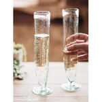 Two Recycled Tall Champagne Flute glasses with sparkling beverage, one being held, showcasing elegant design for dining and entertaining decor from Knotty and Board. 1