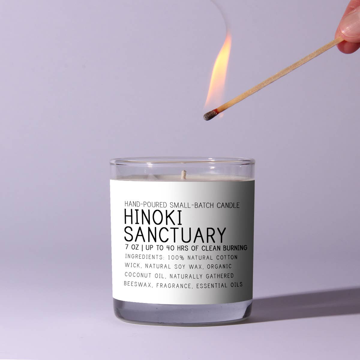 A hand lighting the Just Bee Hinoki Sanctuary Candle, 7 oz. in a clear glass jar with a minimalist white label, perfect for decor and gifts from Knotty and Board.