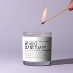 A hand lighting the Just Bee Hinoki Sanctuary Candle, 7 oz. in a clear glass jar with a minimalist white label, perfect for decor and gifts from Knotty and Board. 2