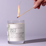Hand lighting a Just Bee White Birch Candle, 13 oz in a clear glass jar with minimalist white and green label, ideal for decor and gifts by Knotty and Board. 1