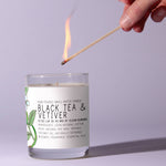 Hand lighting a Just Bee Black Tea Vetiver, 13 oz candle in a clear glass jar with a botanical label, perfect for cozy decor and unique gift ideas from Knotty and Board. 1