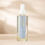 Clear glass bottle of Rockaway Beach Room Spray with a white spray nozzle, featuring a blue label with gold and white text, set against a soft beige background. 1