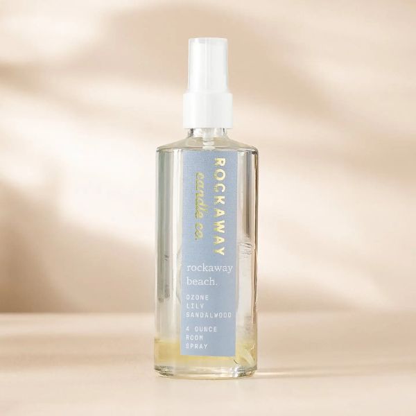 Clear glass bottle of Rockaway Beach Room Spray with a white spray nozzle, featuring a blue label with gold and white text, set against a soft beige background.