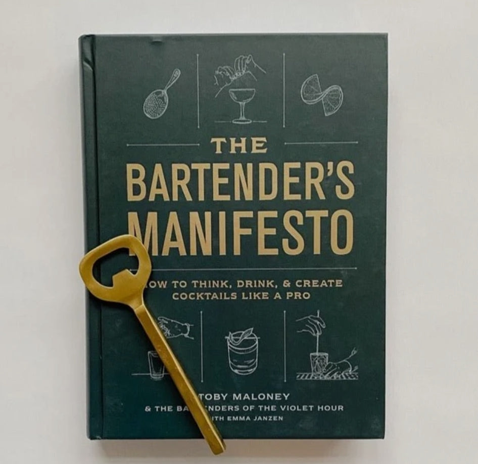 The Bartender's Manifesto book with a dark cover featuring cocktail illustrations, paired with a gold bottle opener, ideal for kitchen decor and entertaining gifts from Knotty and Board.