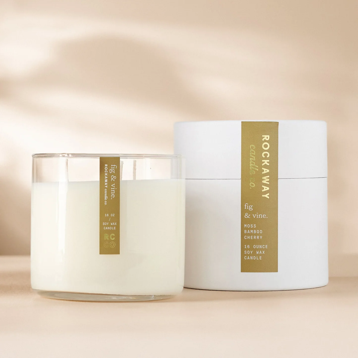 Rockaway Fig & Vine Candle 16oz in a clear glass container with white wax beside its elegant white cylindrical packaging, styled as curated decor and gift from Knotty and Board.